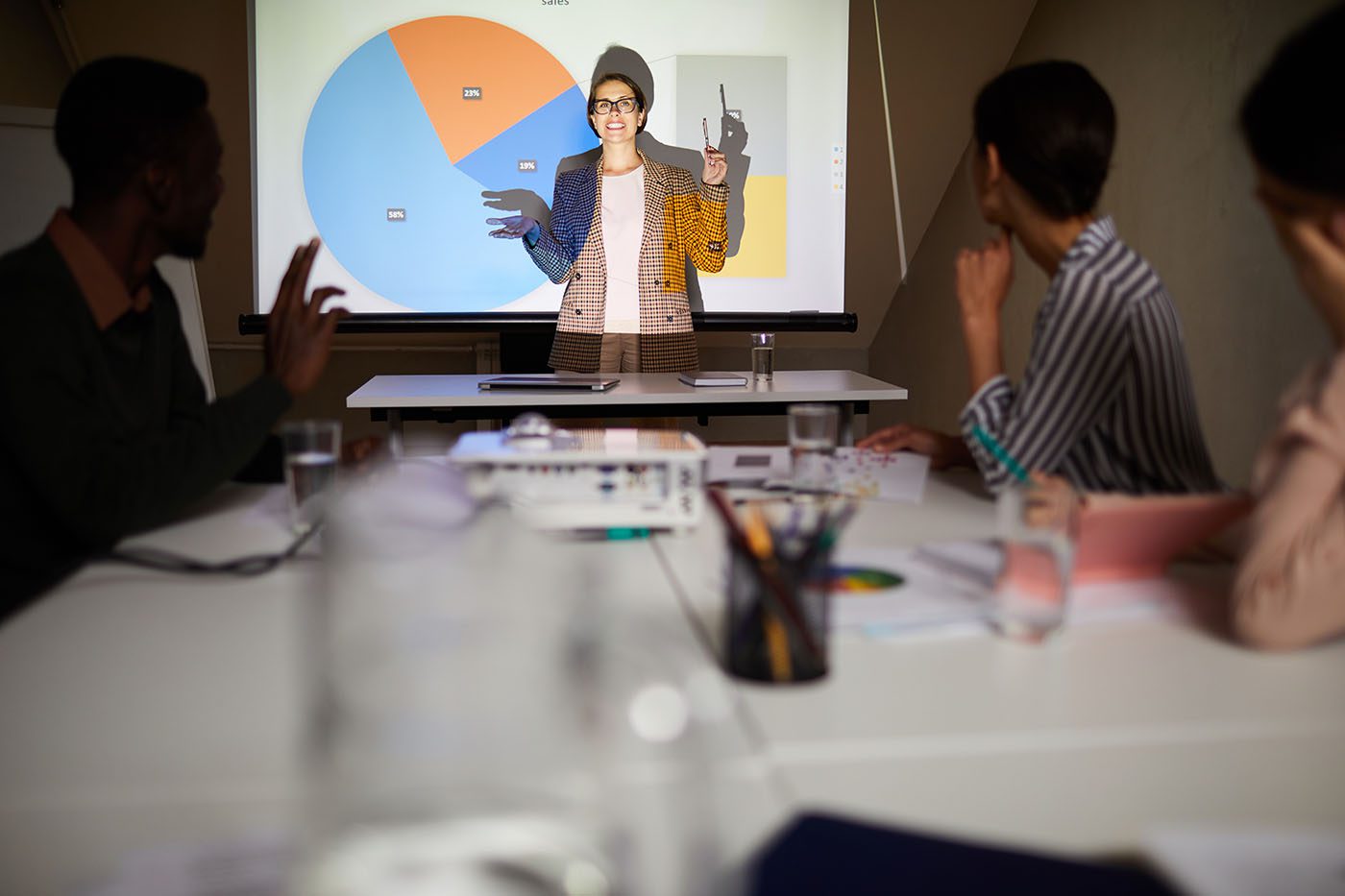 Positive confident female financial analyst in jacket standing against projector and presenting charts in front of audience at meeting